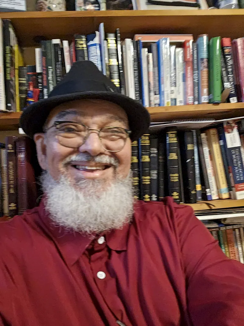 Francisco Brandão, o autor, sorrindo diante de uma estante de livros, de chapéu preto, óculos, barba branca e camisa vinho