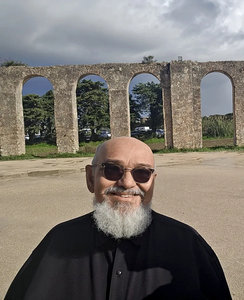 Francisco Brandão diante de um aqueduto antigo de pedra em Portugal, de óculos escuros e roupa preta