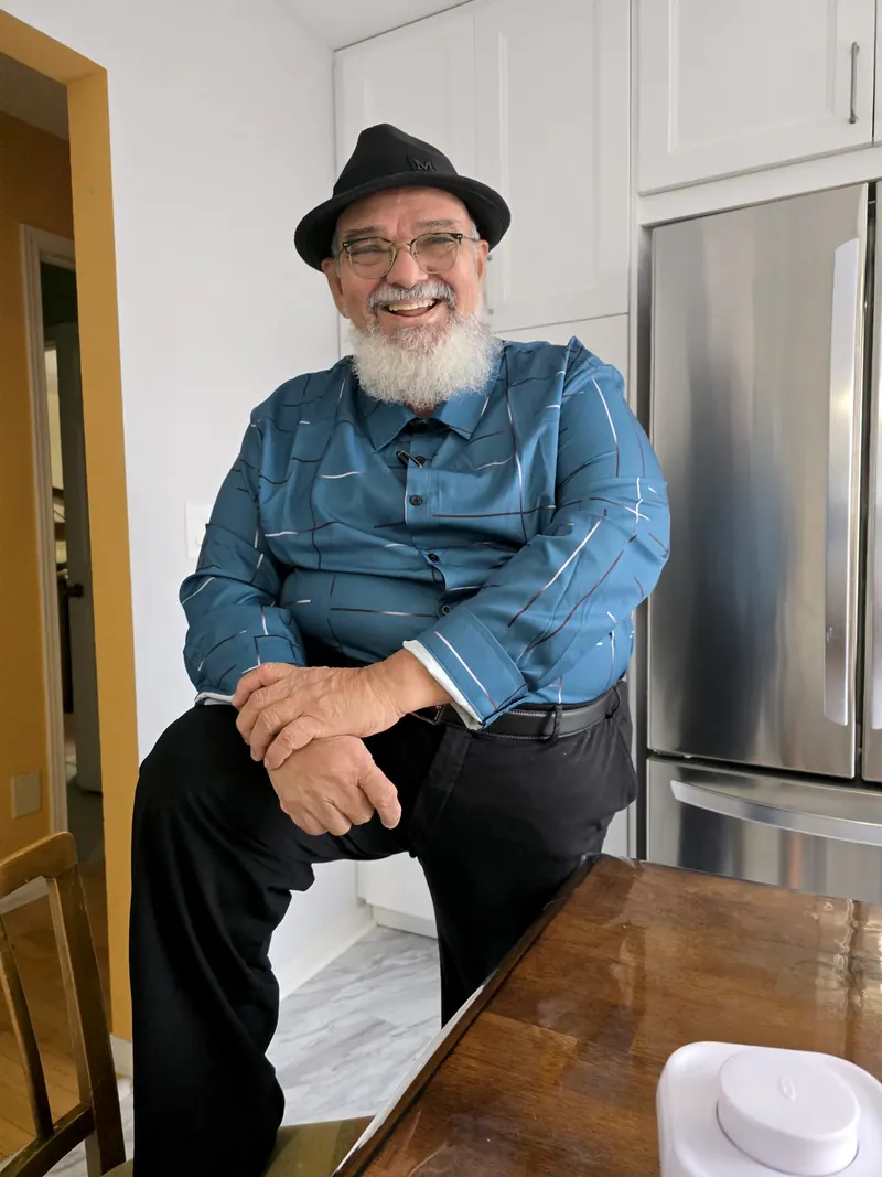 Francisco Brandão sentado, de chapéu, camisa azul e calça preta, sorrindo